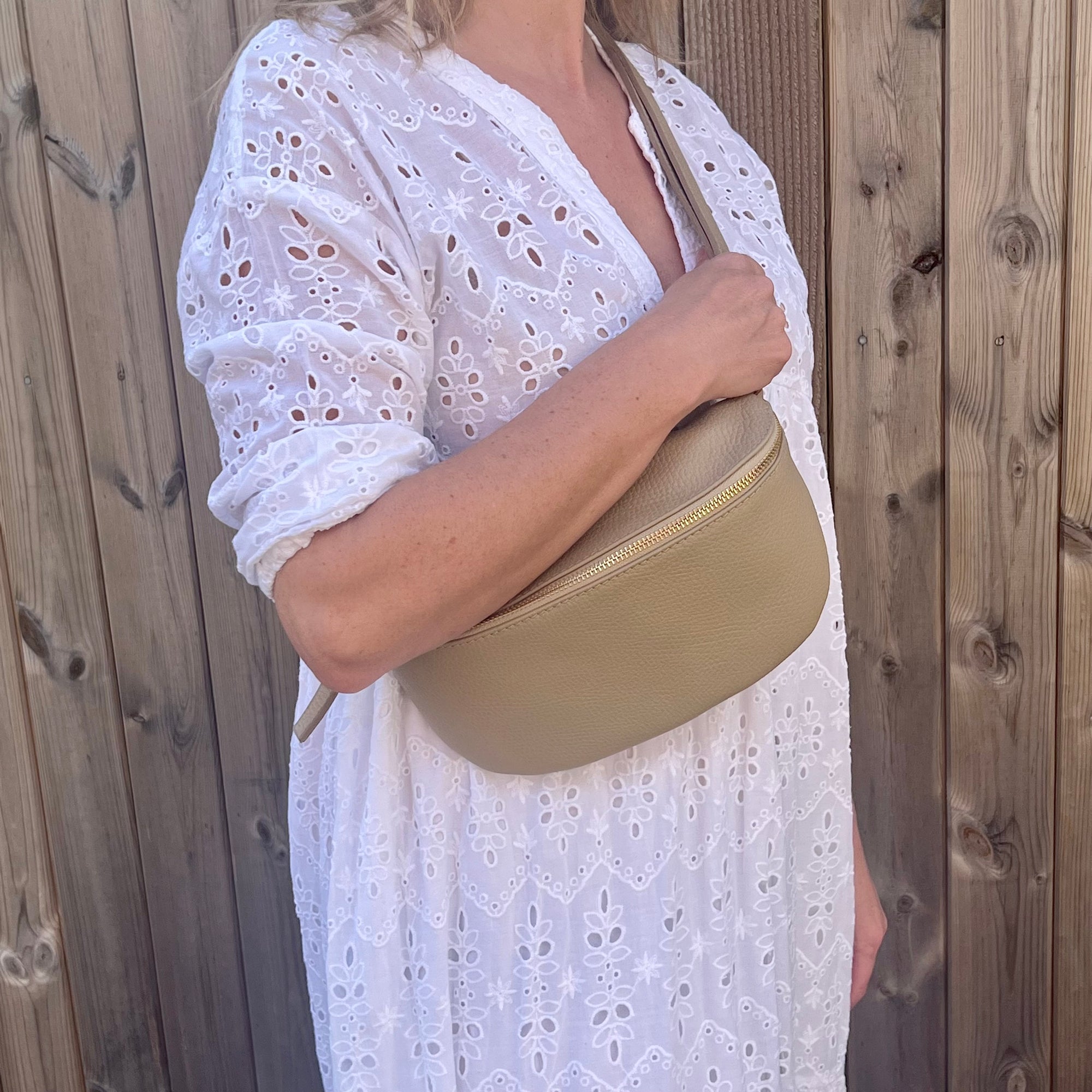 woman wearing a large light taupe crossbody/ waist bag  with and adjustable strap and gold hardware. 
