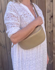 woman wearing a large light taupe crossbody/ waist bag  with and adjustable strap and gold hardware. 
