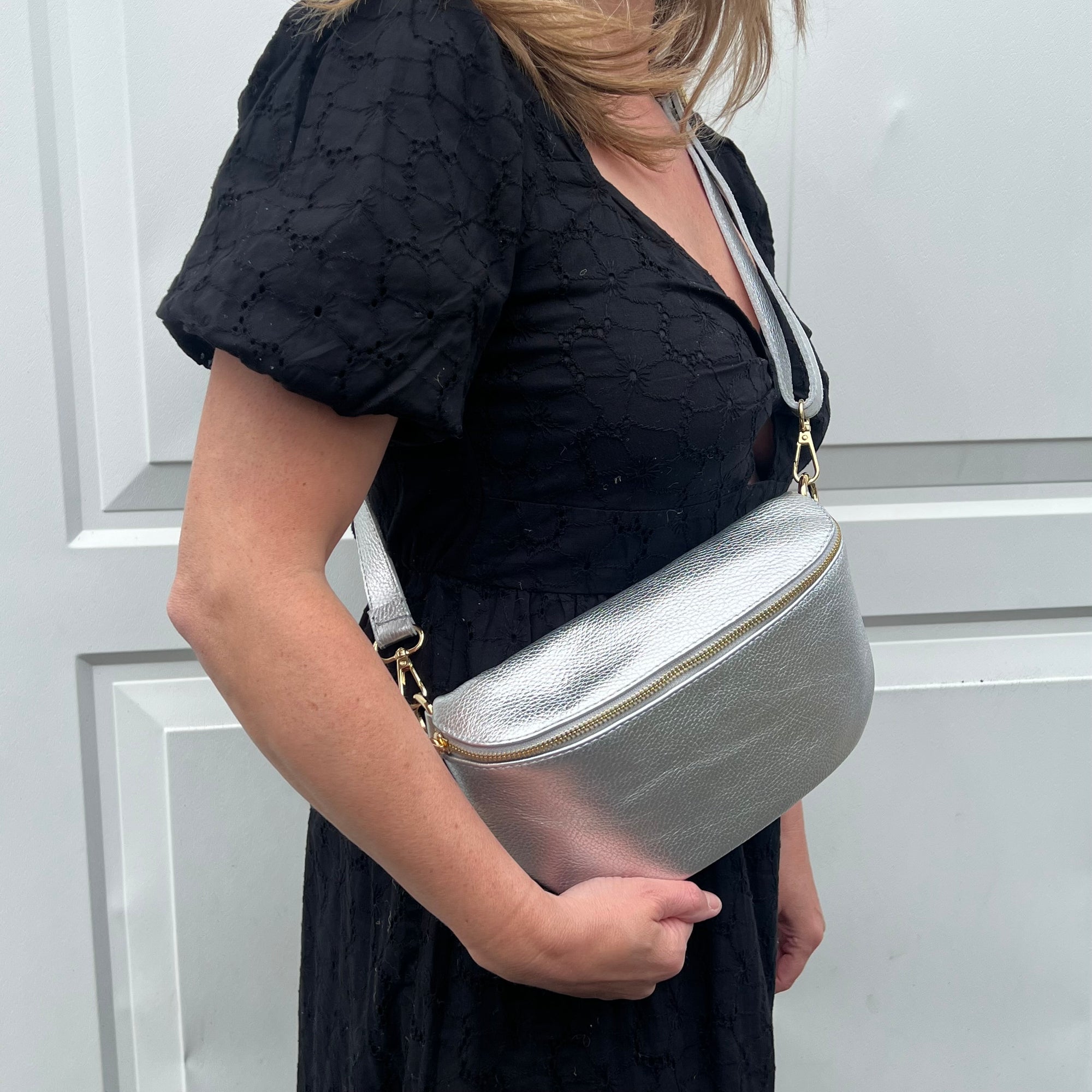 A woman wearing a black dress is holding a large silver leather crossbody bag with an adjustable strap.
