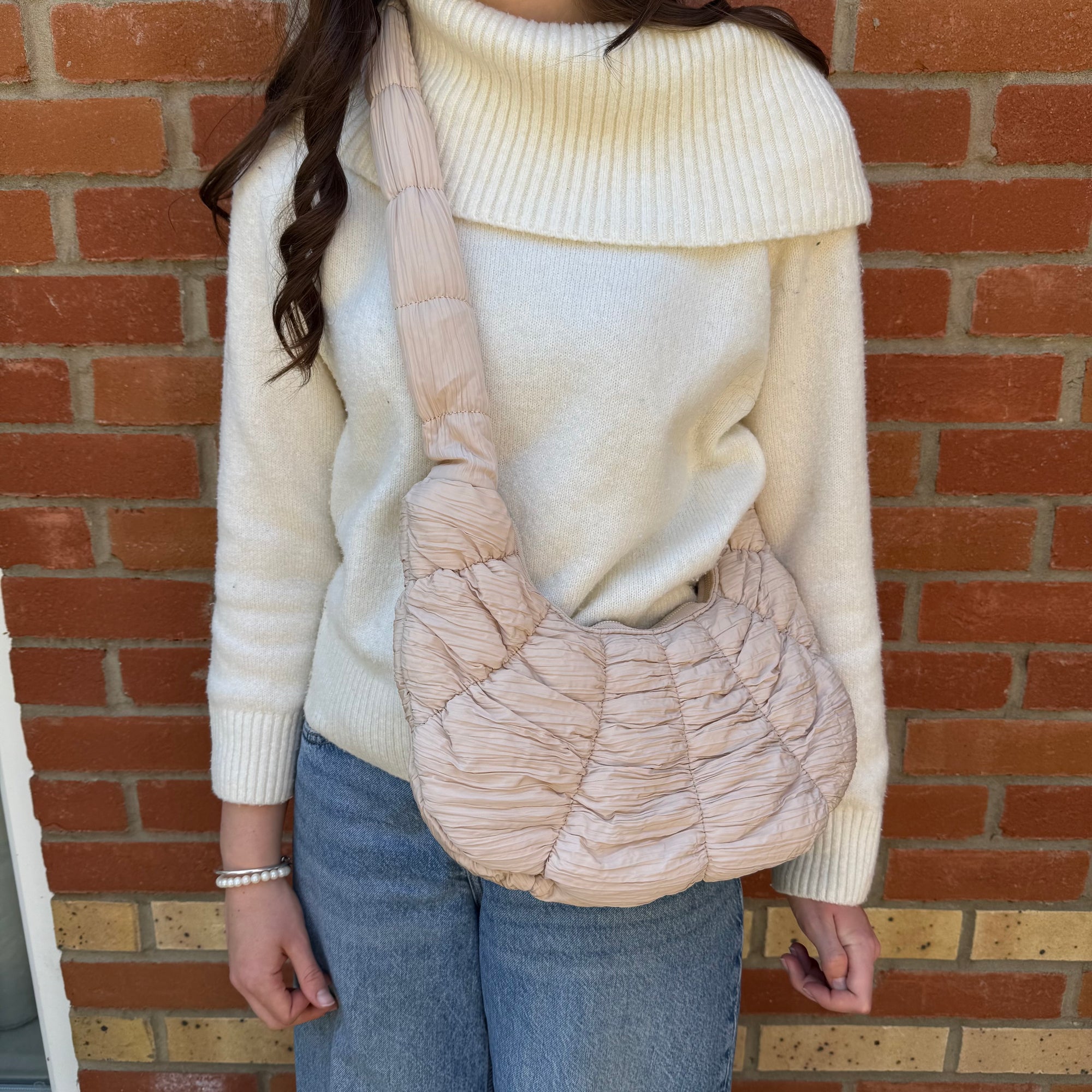 A woman wearing a beige turtleneck sweater holding a latte quilted fabric croissant shoulder bag with a zipper top and gold hardware.