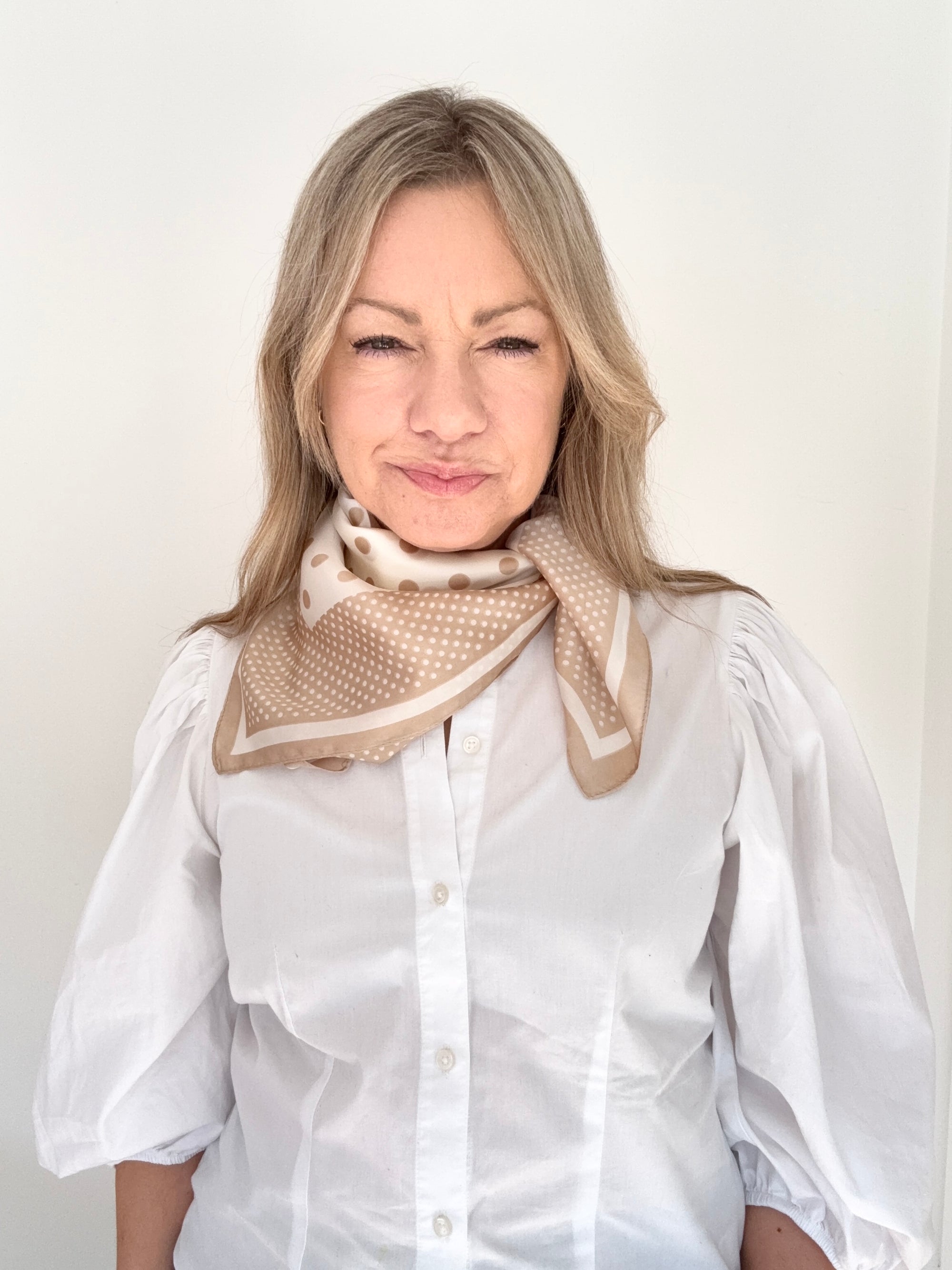 Woman wearing a white shirt and a light camel and cream patterned scarf against a plain background.