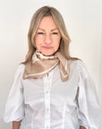 Woman wearing a white shirt and a light camel and cream patterned scarf against a plain background.