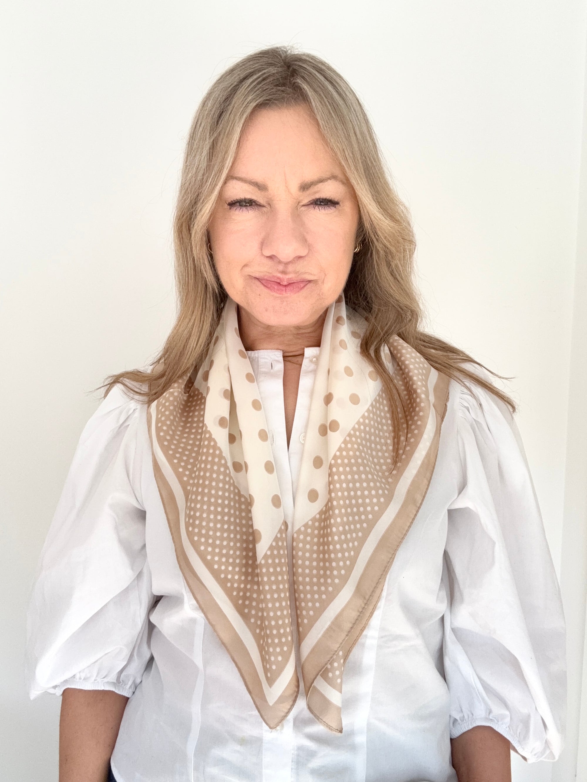 Woman wearing a white shirt and a light camel and cream patterned scarf against a plain background.