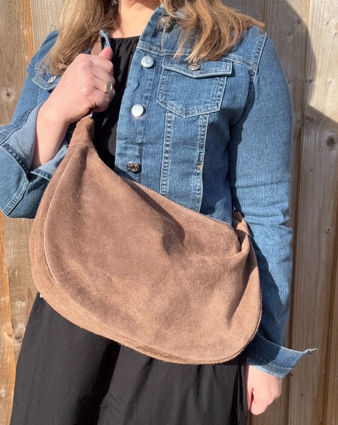 A woman wearing a denim jacket holding a mocha brown suede large crossbody swing bag with a detachable strap.