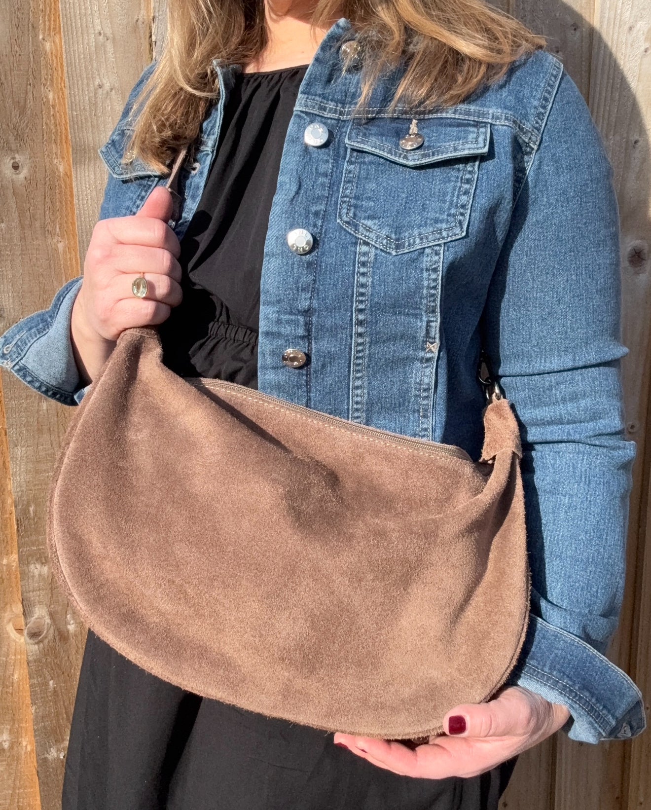 A woman wearing a denim jacket holding a mocha brown suede large crossbody swing bag with a detachable strap.