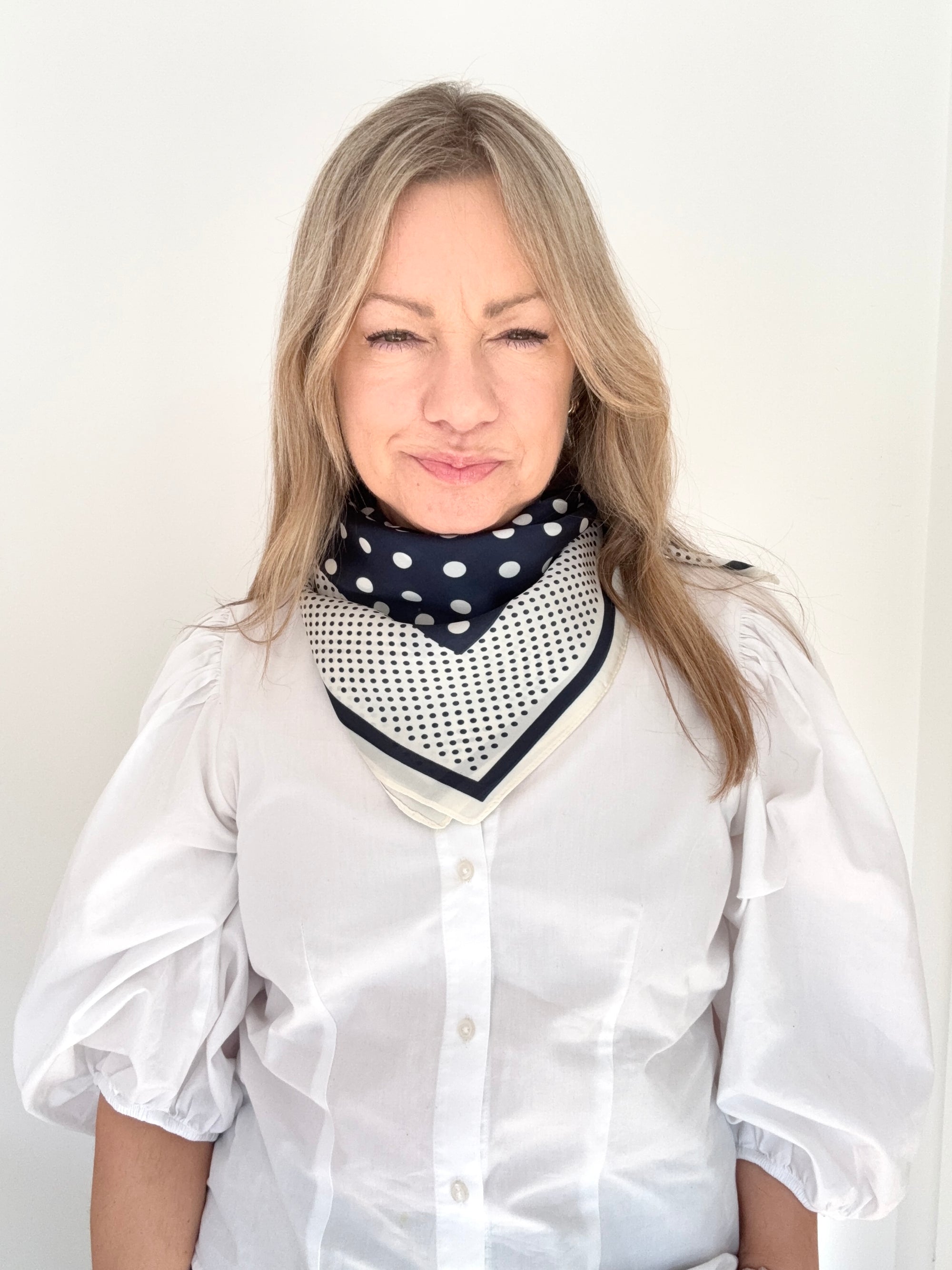 Woman wearing a white shirt and navy and cream patterned scarf against a plain background.