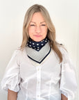 Woman wearing a white shirt and navy and cream patterned scarf against a plain background.