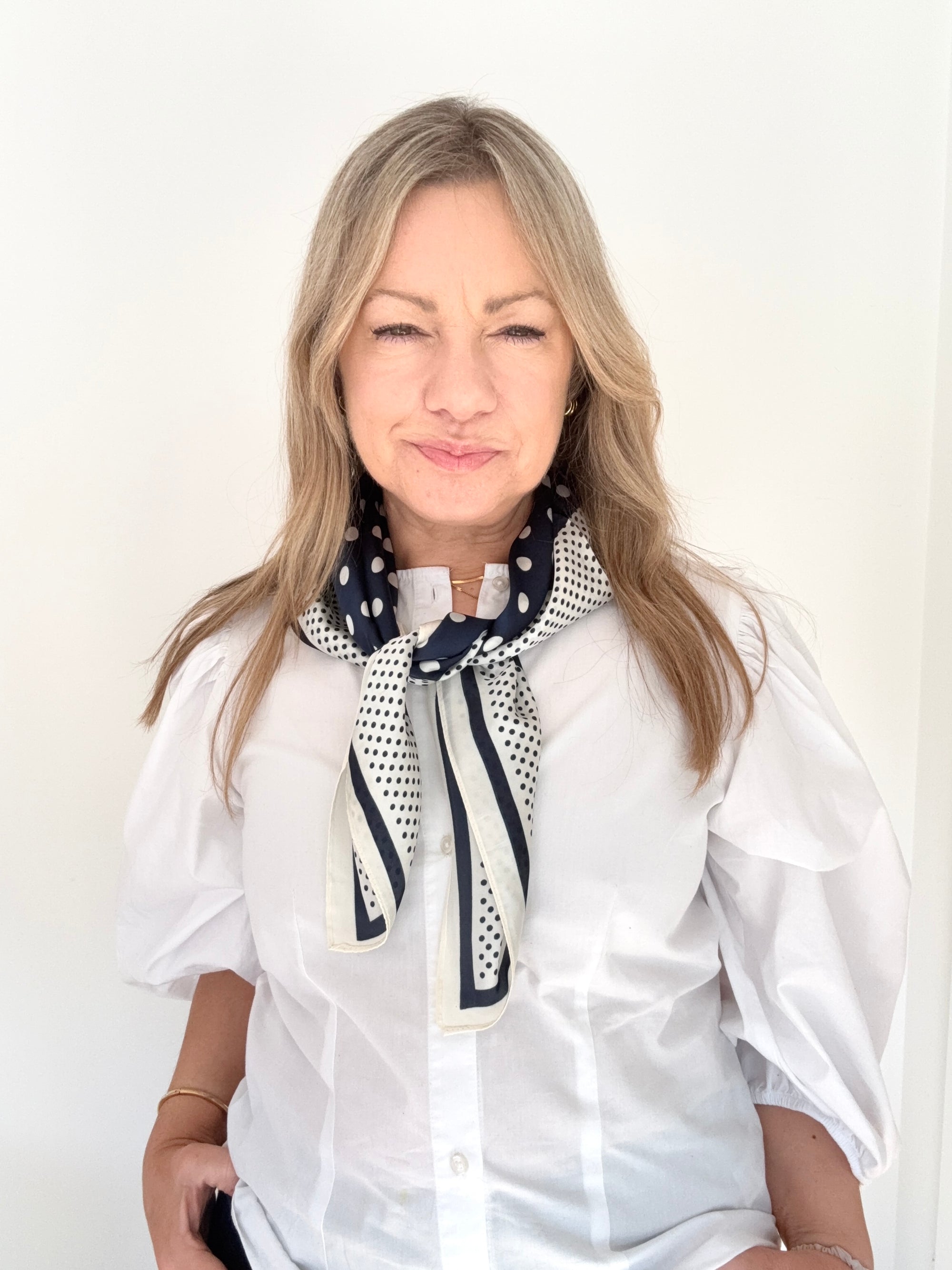 Woman wearing a white shirt and navy and cream patterned scarf against a plain background.