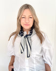 Woman wearing a white shirt and navy and cream patterned scarf against a plain background.