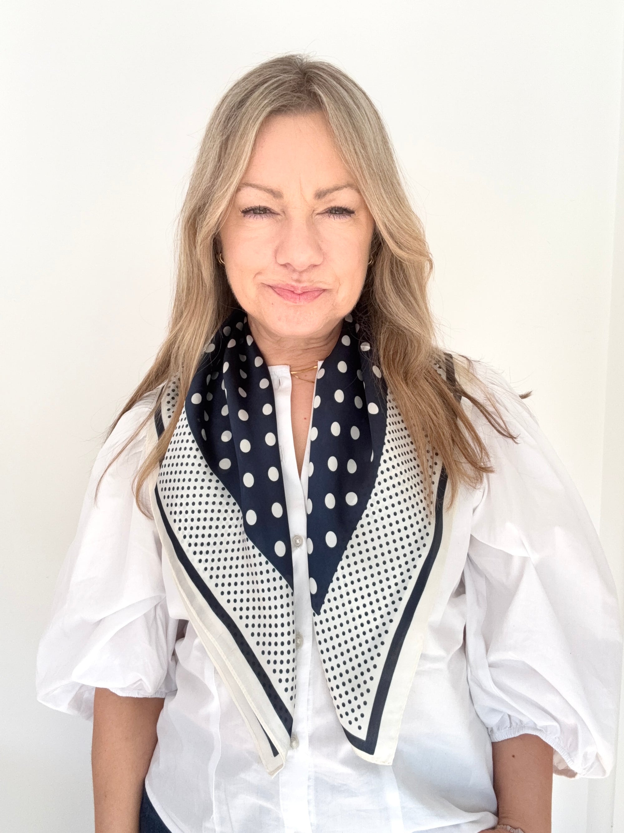 Woman wearing a white shirt and navy and cream patterned scarf against a plain background.