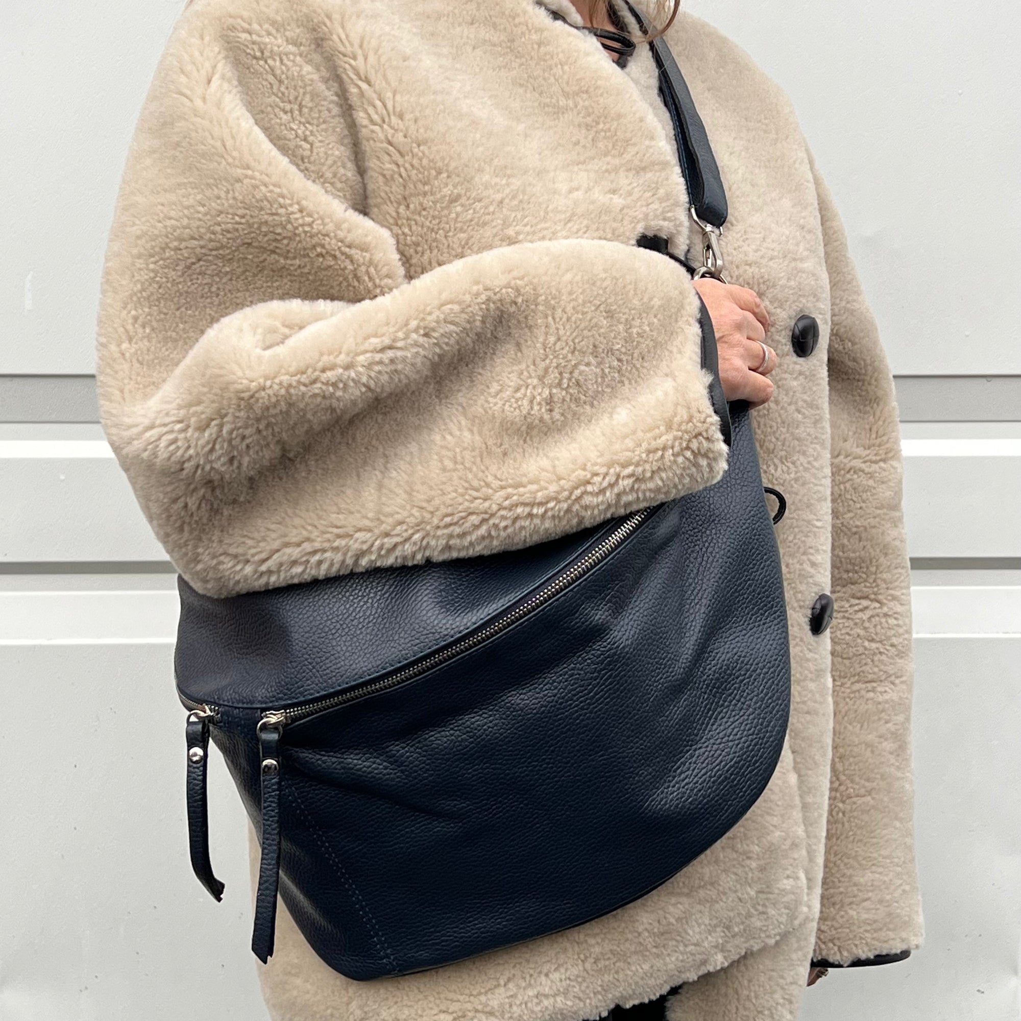 A woman wearing a beige coat is holding a navy blue leather crossbody bag with an adjustable strap and silver hardware.
