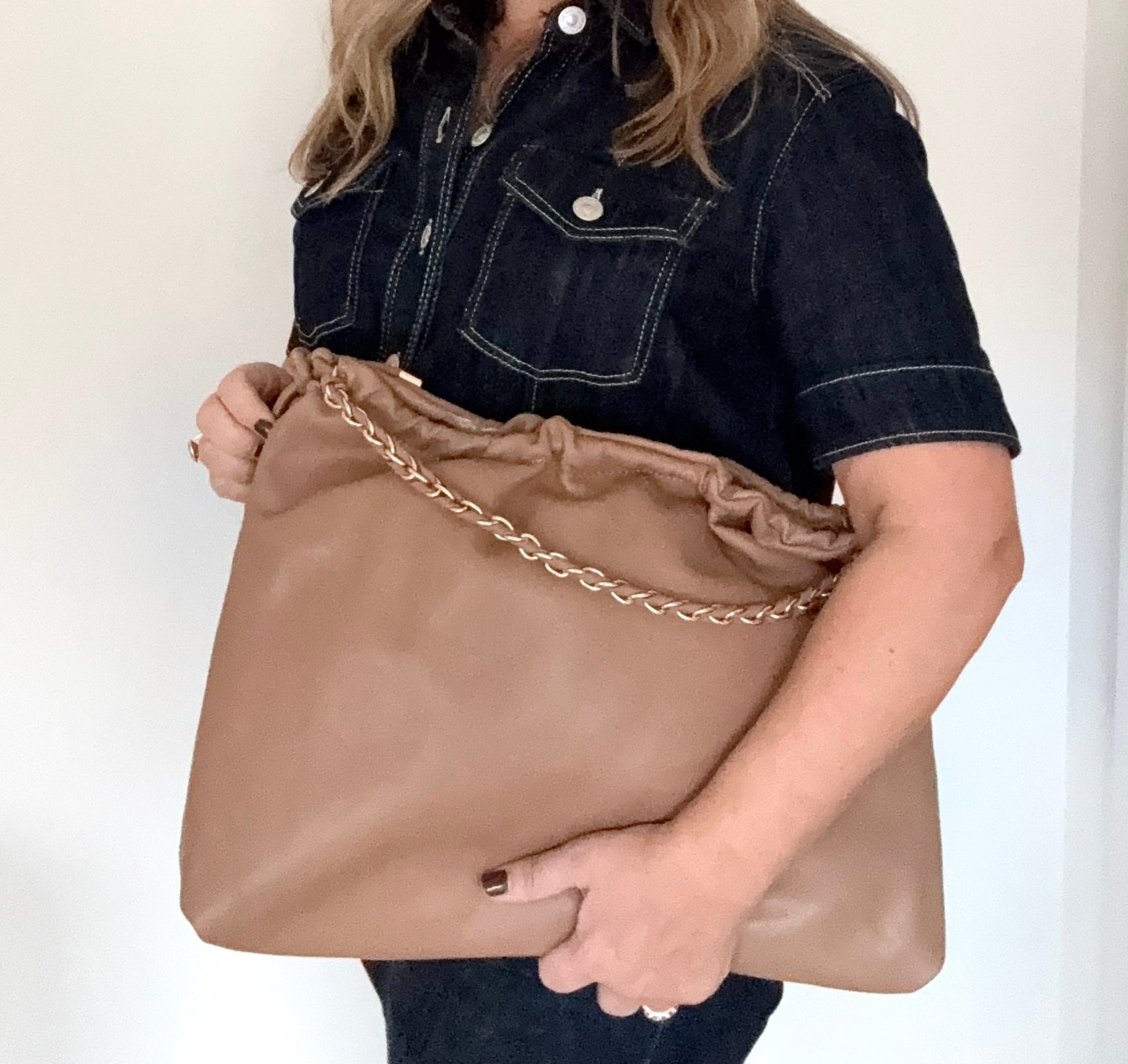 Woman holding a pale camel faux  leather handbag with a gold chain strap against 