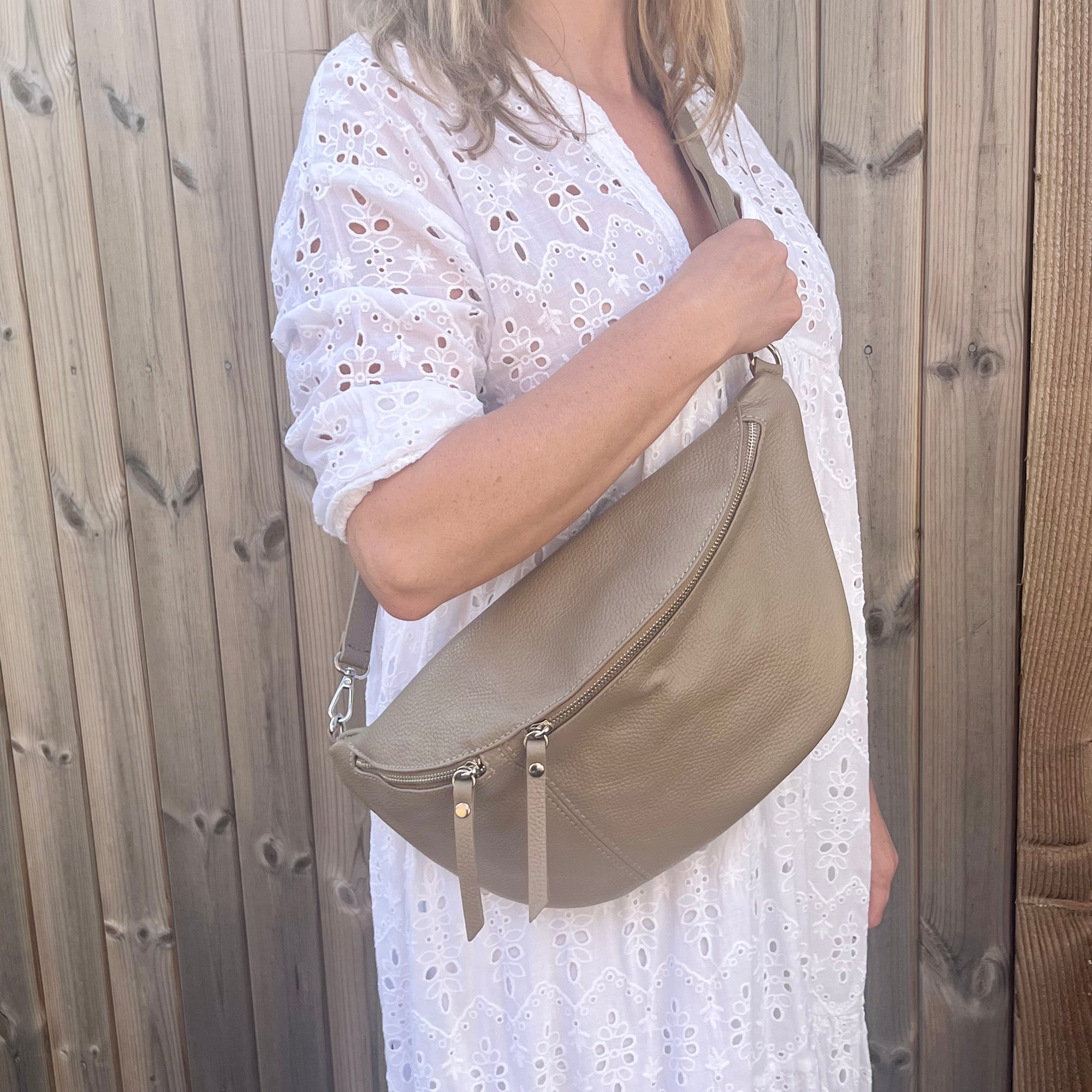 A person wearing a white dress is holding a pale taupe large crossbody bum bag with an adjustable, detachable leather strap and silver hardware.