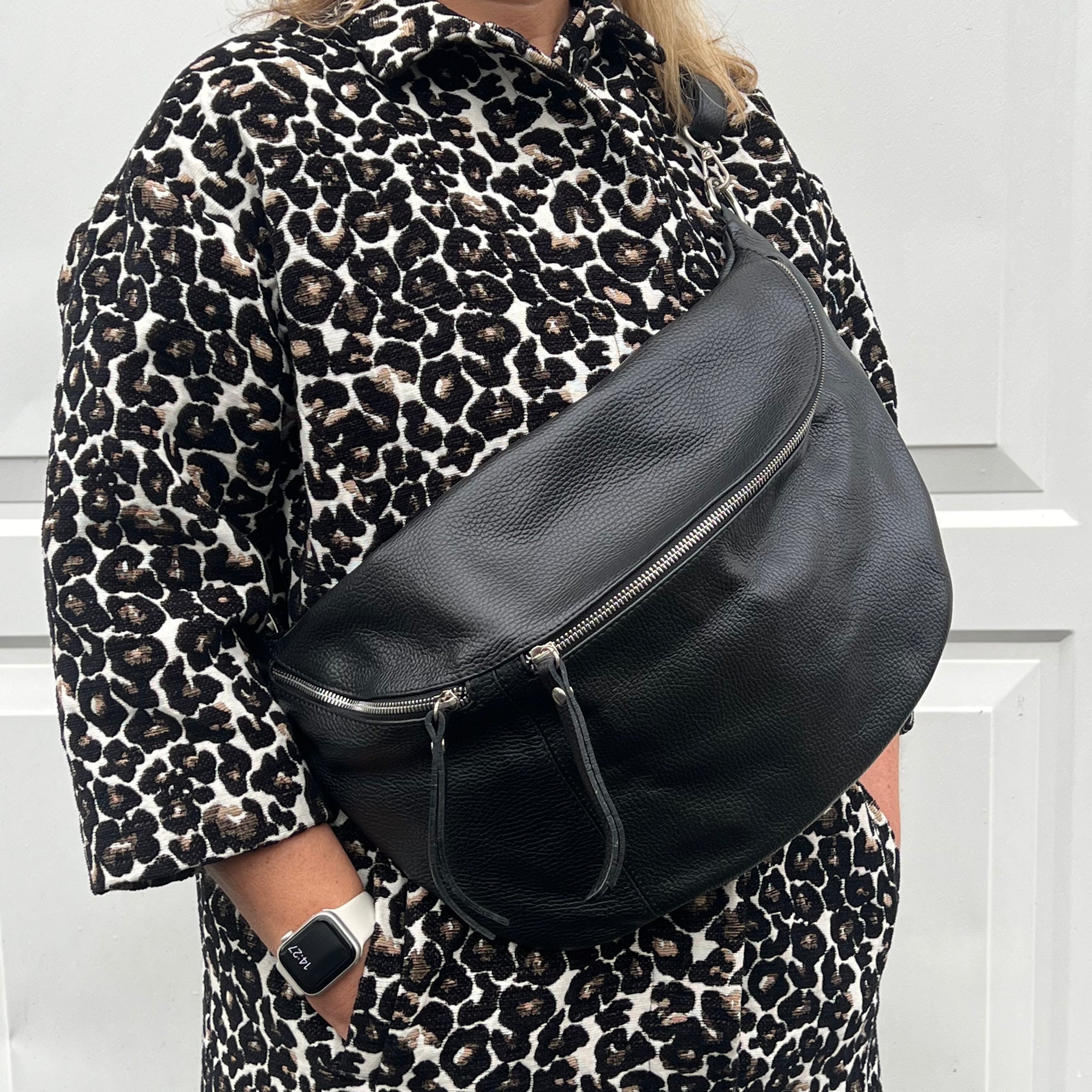 Woman wearing a black leather crossbody bag with silver hardware and an adjustable strap over a leopard print coat.