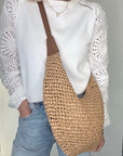 Woman wearing a white top, blue jeans, holding a raffia and tan woven crossbody handbag.