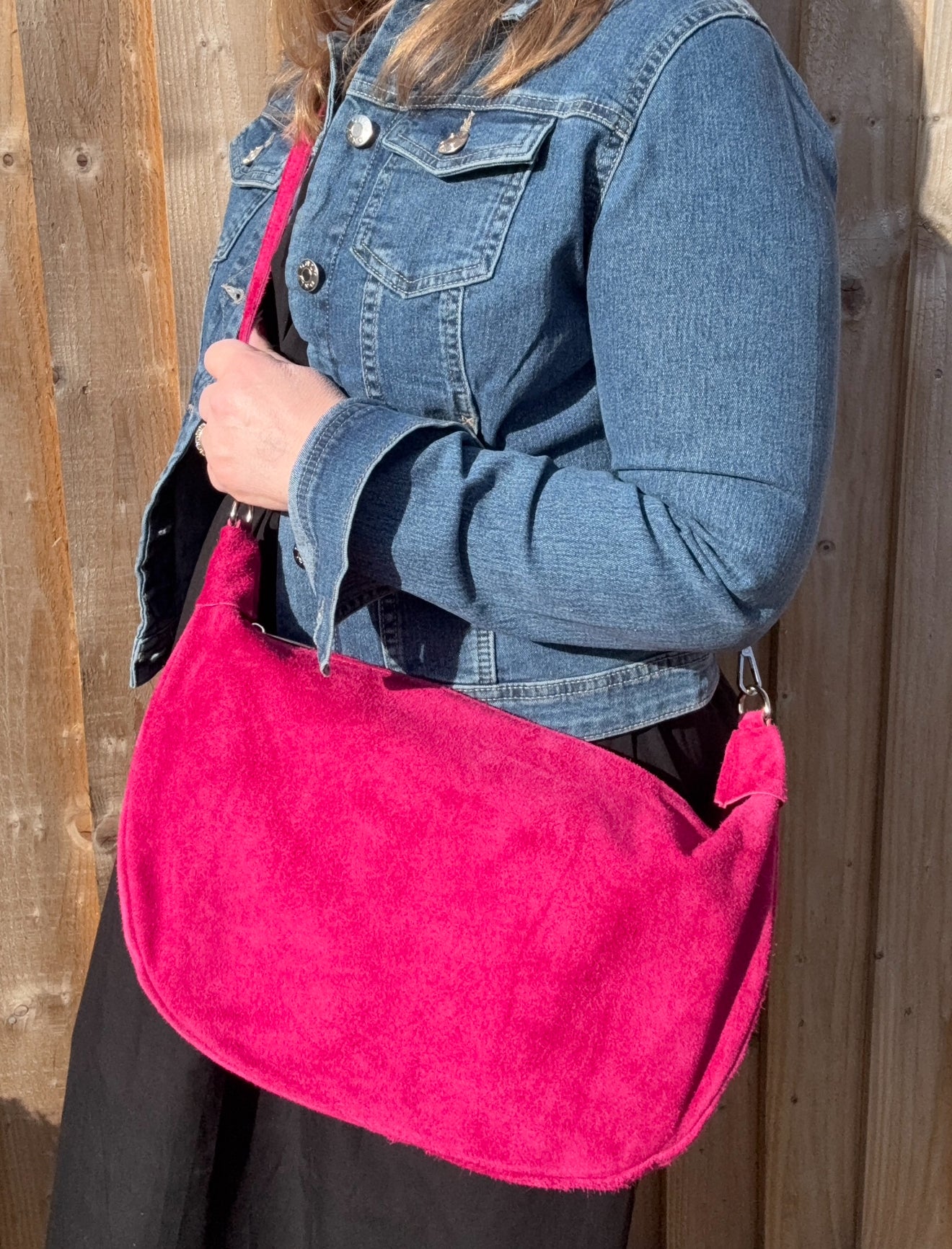 A person wearing a denim jacket is holding a raspberry  large suede leather crossbody swing bag with a detachable strap.