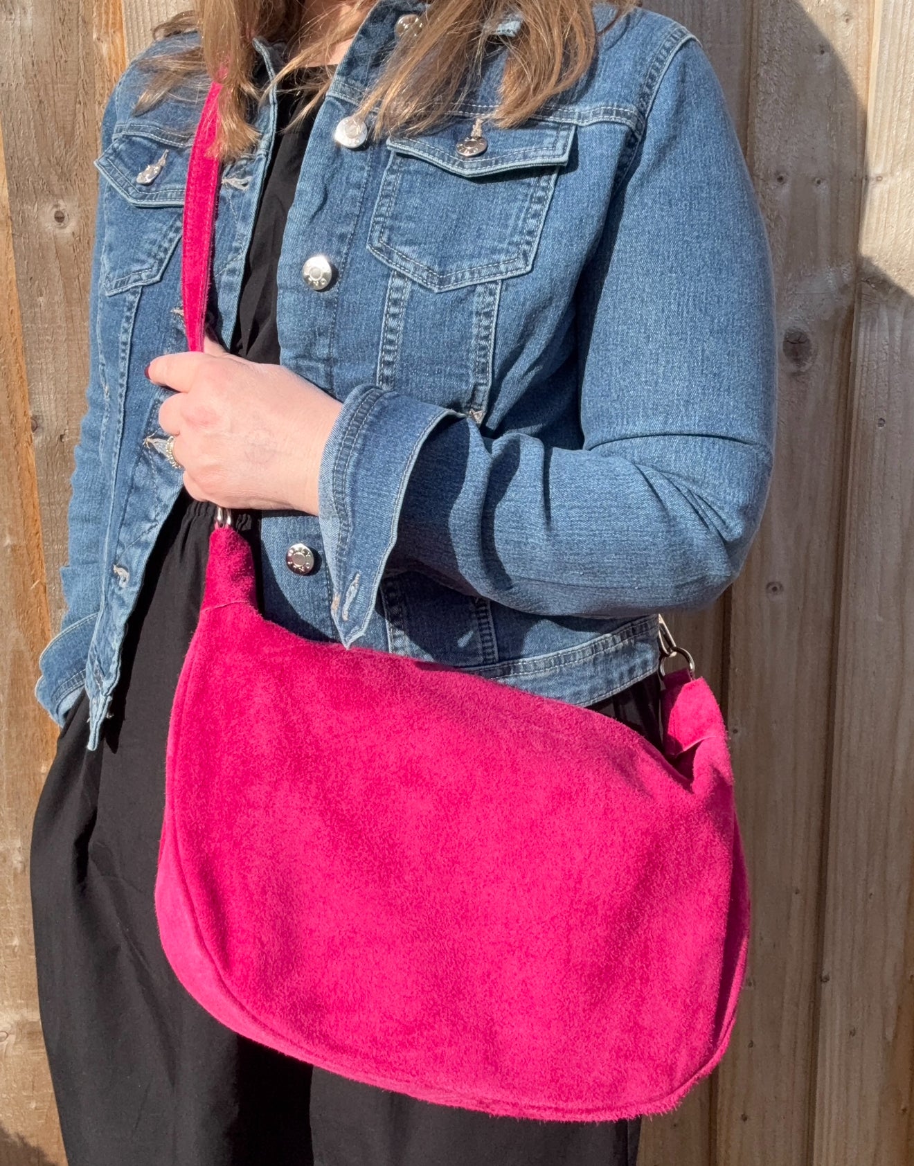 A person wearing a denim jacket is holding a raspberry  large suede leather crossbody swing bag with a detachable strap.