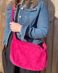 A person wearing a denim jacket is holding a raspberry  large suede leather crossbody swing bag with a detachable strap.
