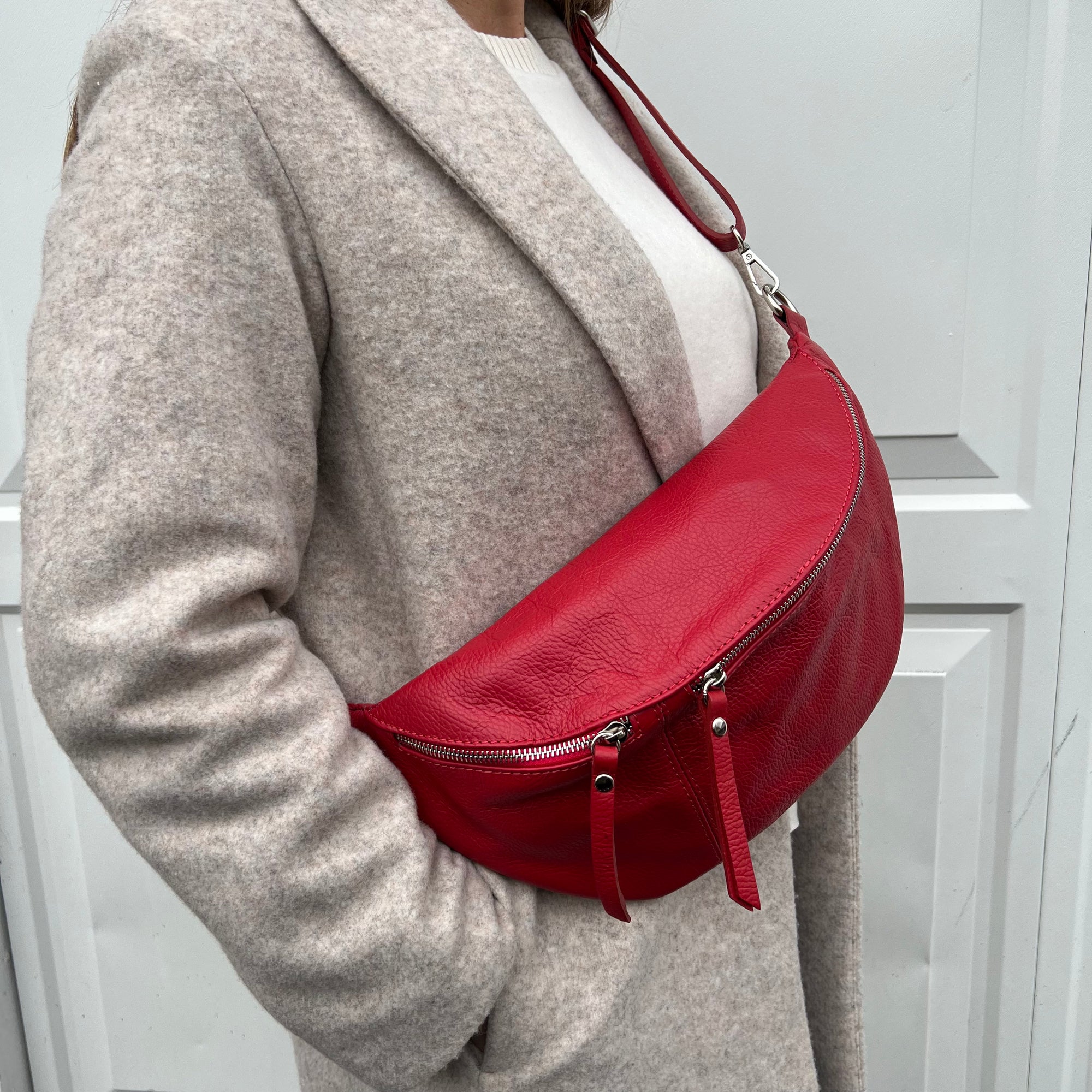 A woman  wearing a grey coat is carrying a red large crossbody bum bag with an adjustable, detachable leather strap and multiple sliver accented zip pockets.