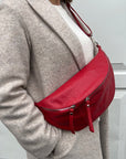 A woman  wearing a grey coat is carrying a red large crossbody bum bag with an adjustable, detachable leather strap and multiple sliver accented zip pockets.