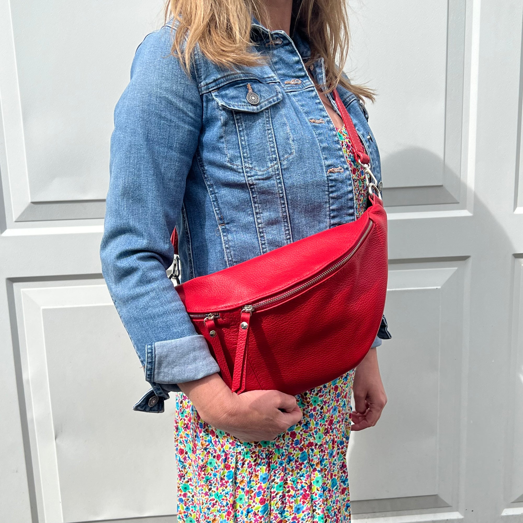 A woman wearing a blue denim jacket and a floral dress, carrying a red large crossbody bum bag with silver hardware.