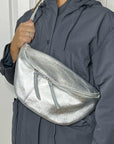 A woman wearing a silver large crossbody bum bag with an adjustable, detachable leather strap and multiple zip pockets.
