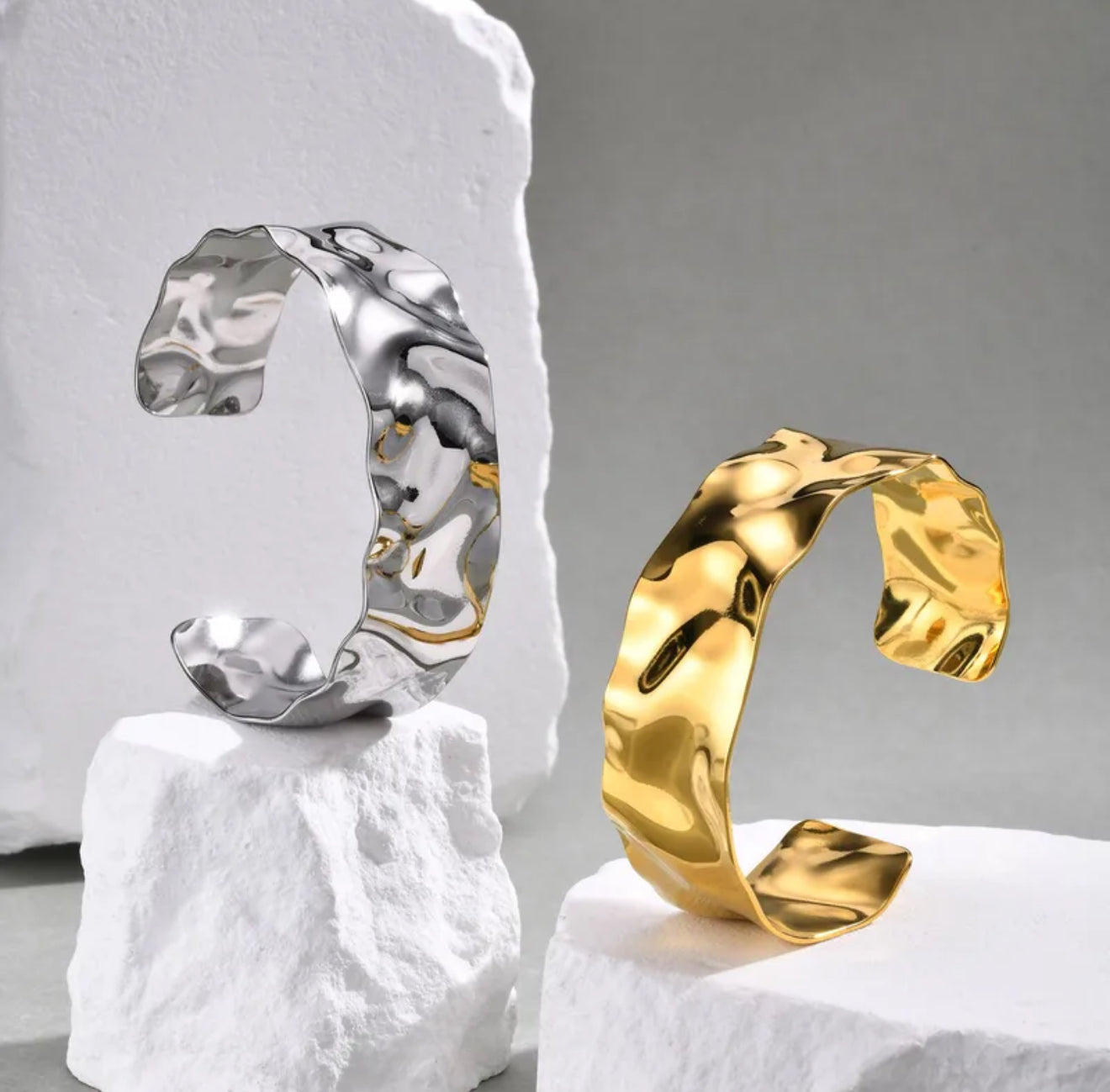 Two unique cuff bangle bracelets, one silver and one gold, on white and grey stone bases.