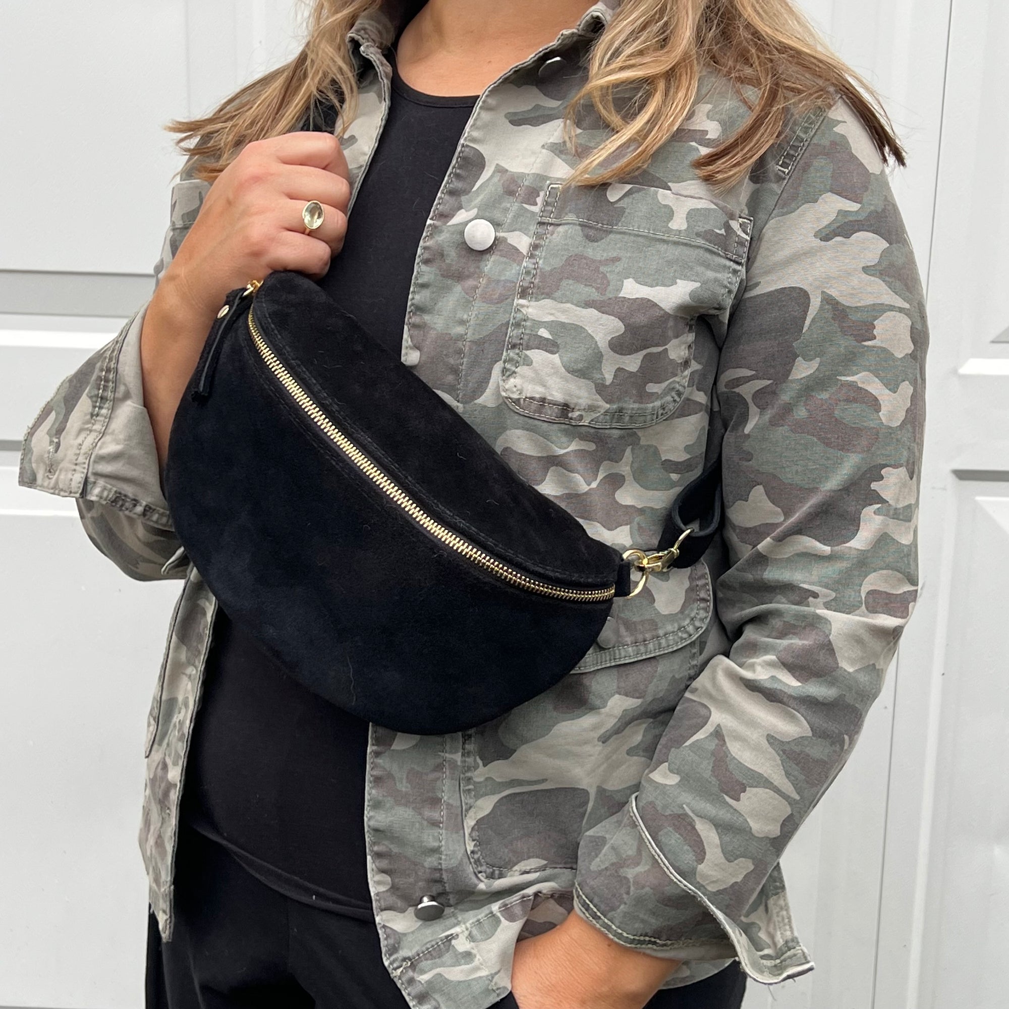 A woman wearing a camouflage jacket holding a black suede crossbody bag with gold-colored hardware.