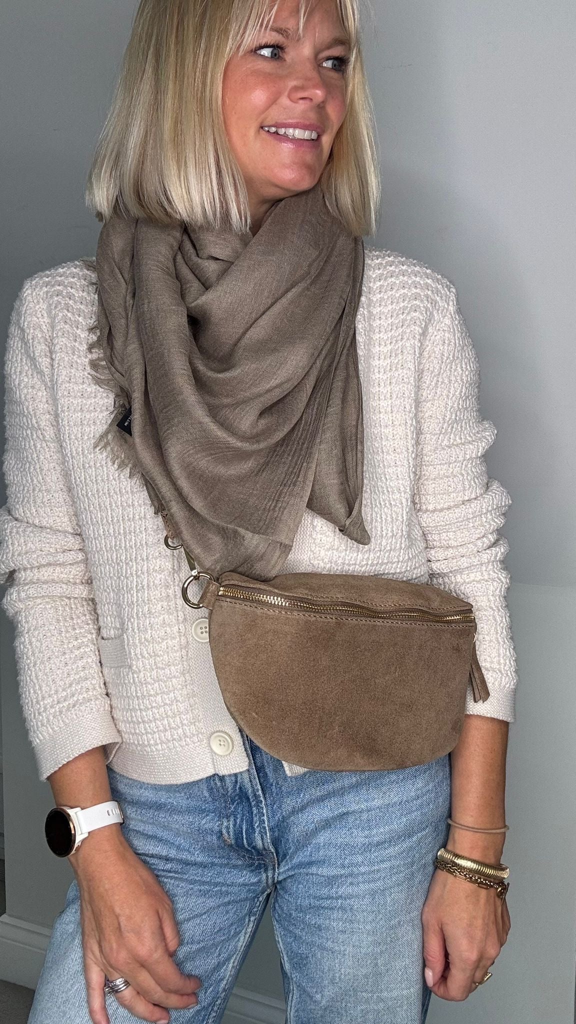 a woman wearing a white jumper and a grey scarf, holding a dark taupe suede crossbody/ waist bag. 