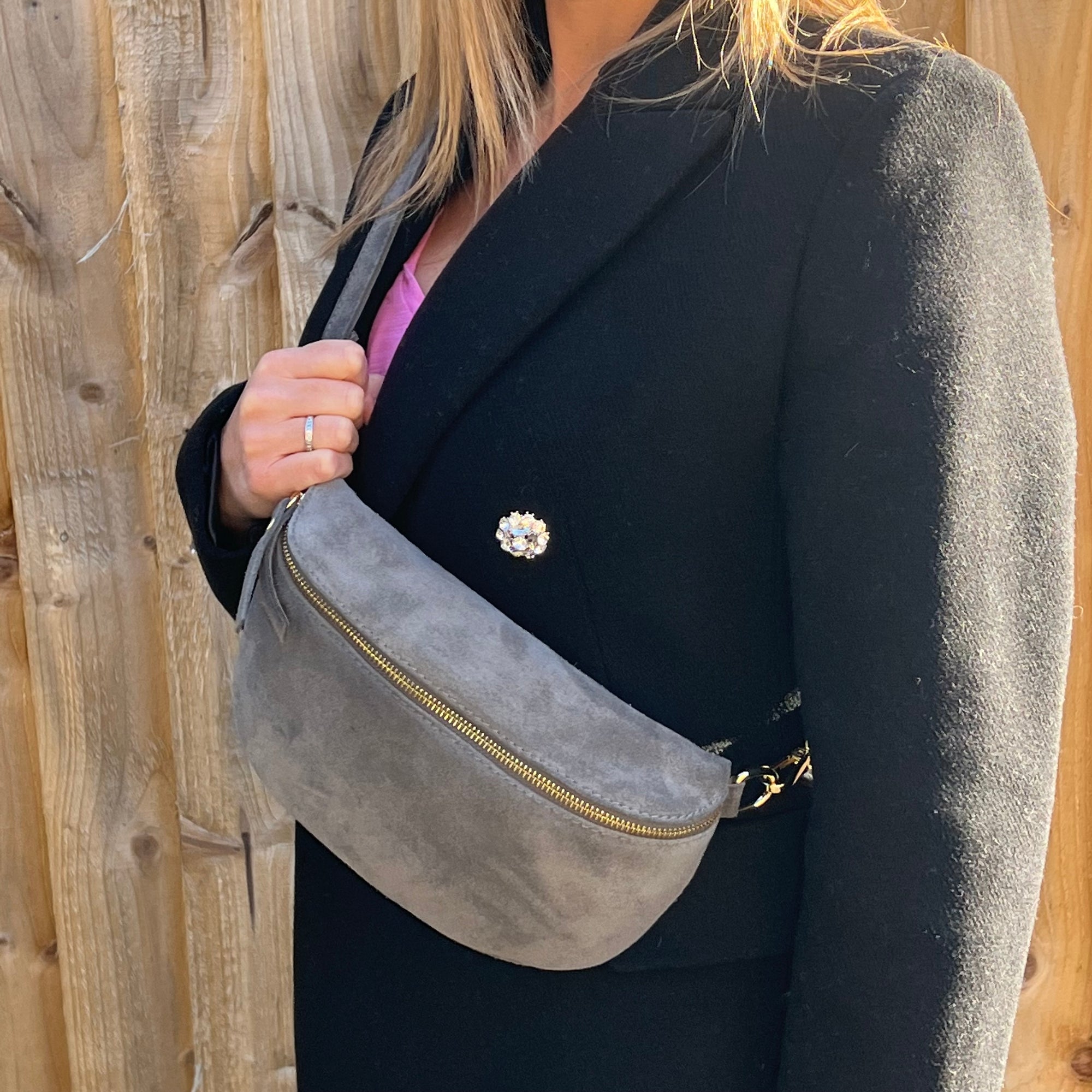 woman wearing a black suede coat holding a suede grey crossbody/ waist bag with gold accents 