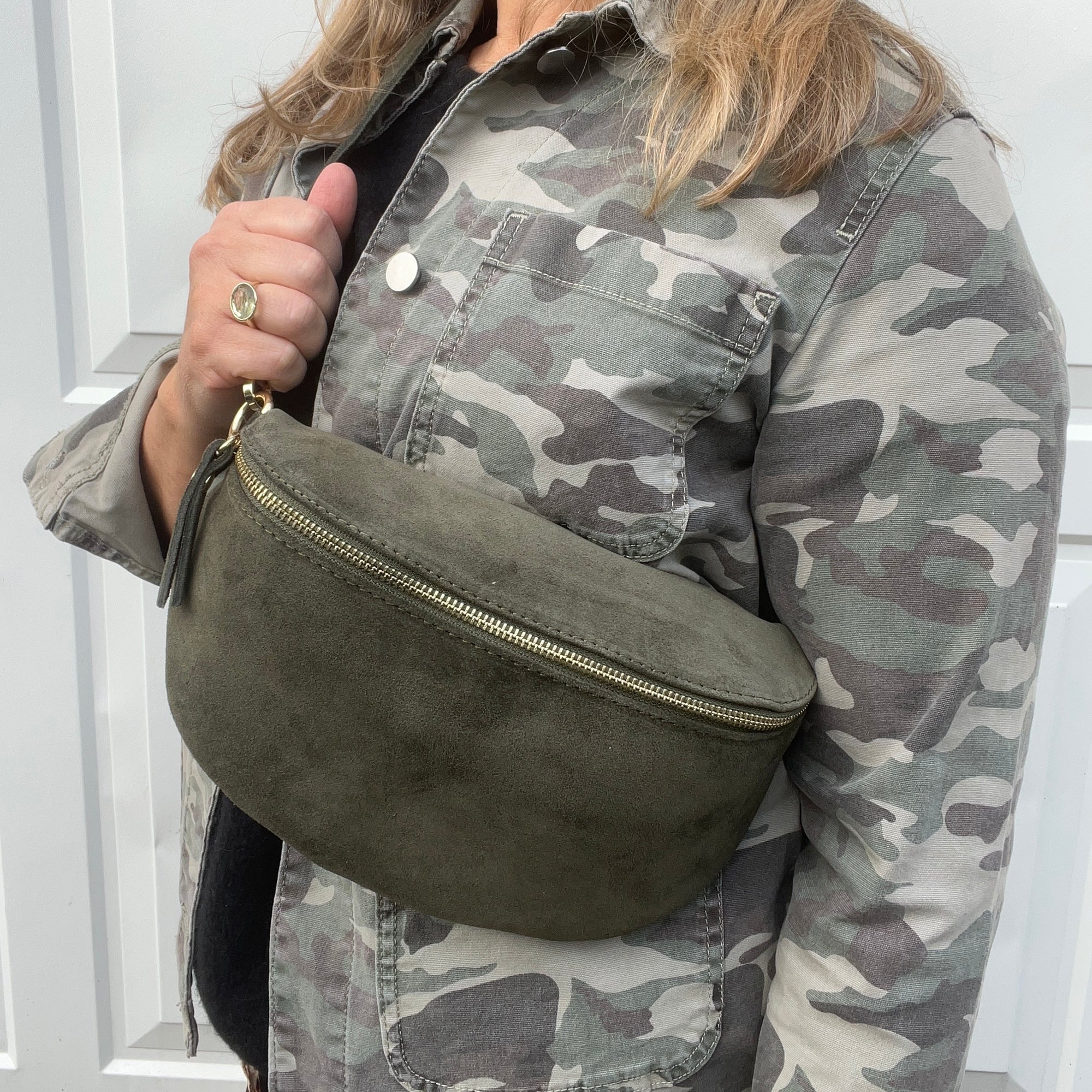 A woman wearing a camouflage jacket holding a khaki suede crossbody bag with gold accents 