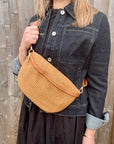 Woman wearing a black dress and denim jacket, holding a woven tan raffia crossbody handbag. 