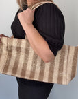 A person holding a taupe and cream raffia tote bag with gold accents and woven thread, over their shoulder.