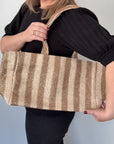 A person holding a taupe and cream raffia tote bag with gold accents and woven thread, over their shoulder.