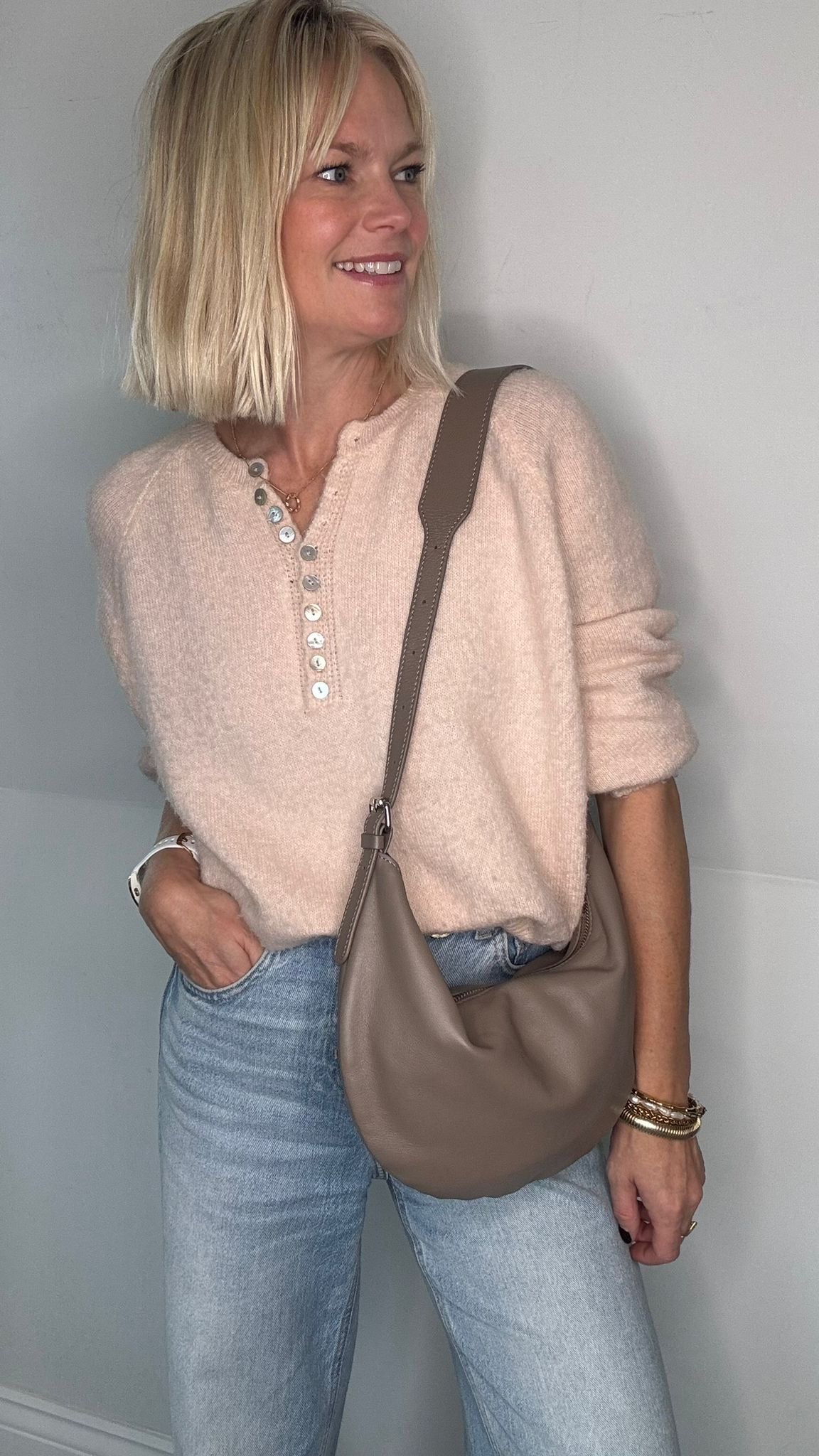 Woman wearing a beige sweater and blue jeans, holding a taupe crossbody/  shoulder bag 