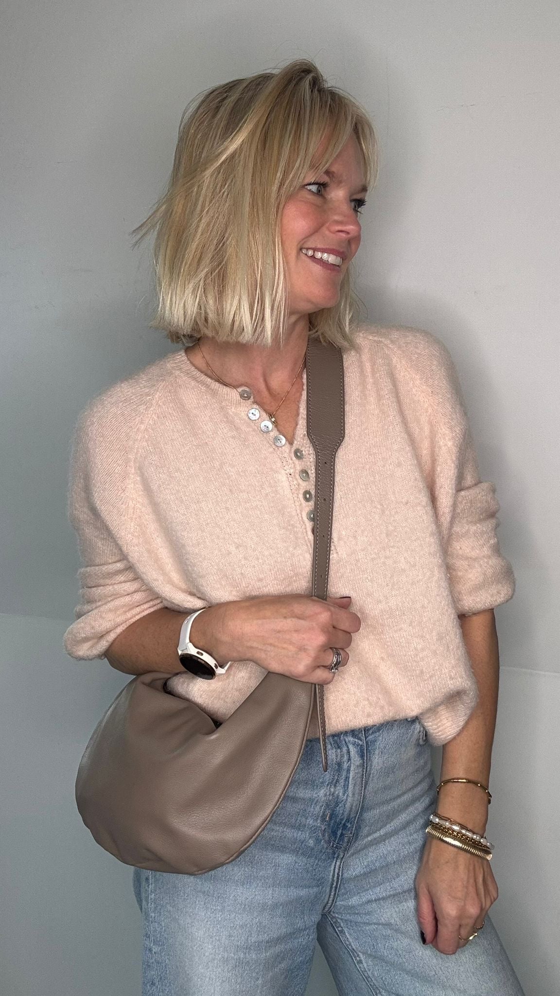Woman wearing a beige sweater and blue jeans, holding a taupe crossbody/  shoulder bag 