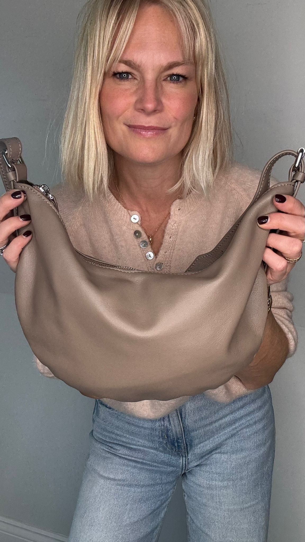 Woman wearing a beige sweater and blue jeans, holding a taupe crossbody/  shoulder bag 