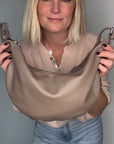 Woman wearing a beige sweater and blue jeans, holding a taupe crossbody/  shoulder bag 