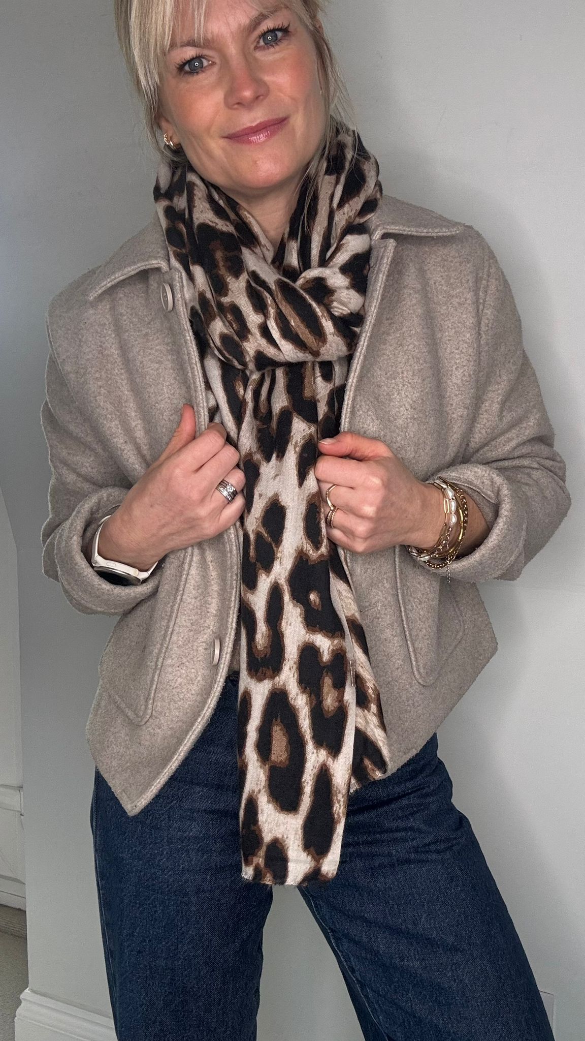 Woman wearing a beige coat, blue jeans, and a  taupe leopard print scarf 