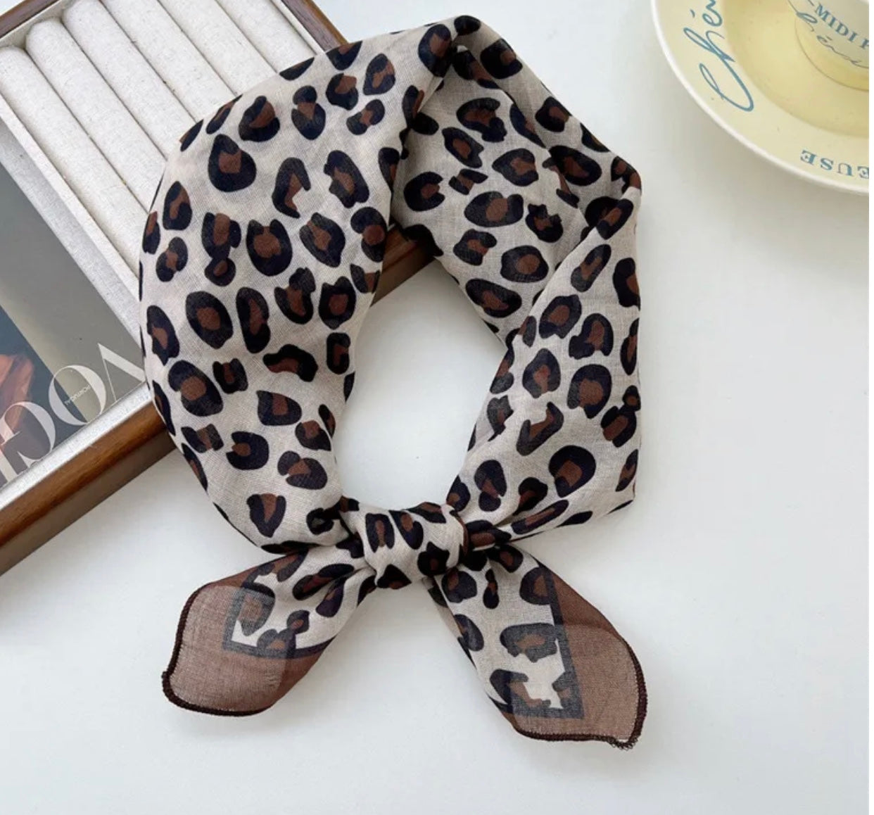 Animal print square scarf on a white surface with a book and plate in the background.