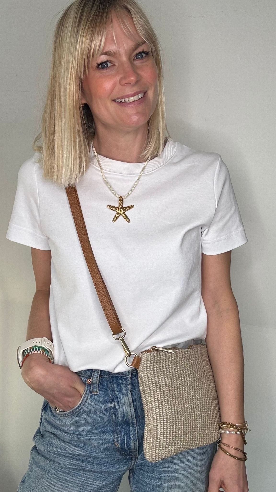 Woman wearing a white t-shirt, blue jeans, and carrying a taupe raffia clutch/ crossbody handbag. 