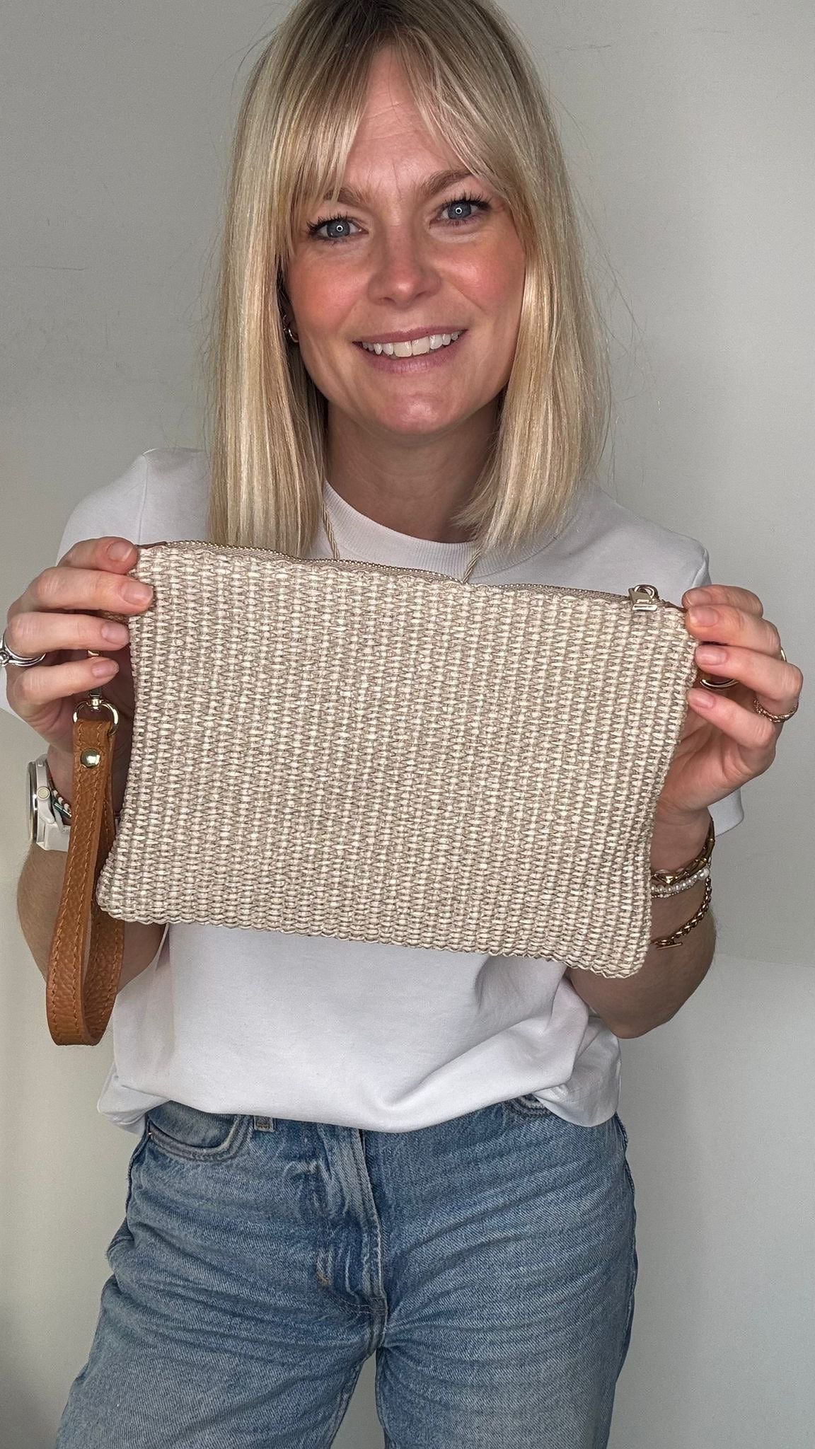 Woman wearing a white t-shirt, blue jeans, and carrying a taupe raffia clutch/ crossbody handbag. 