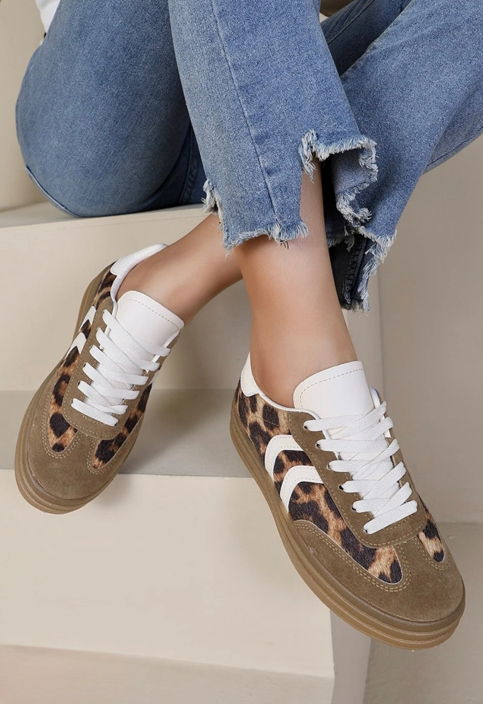 A pair of leopard print wedge lace-up trainers with white detail and laces, worn by a person sitting with jeans on.