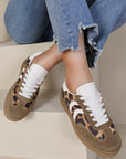 A pair of leopard print wedge lace-up trainers with white detail and laces, worn by a person sitting with jeans on.