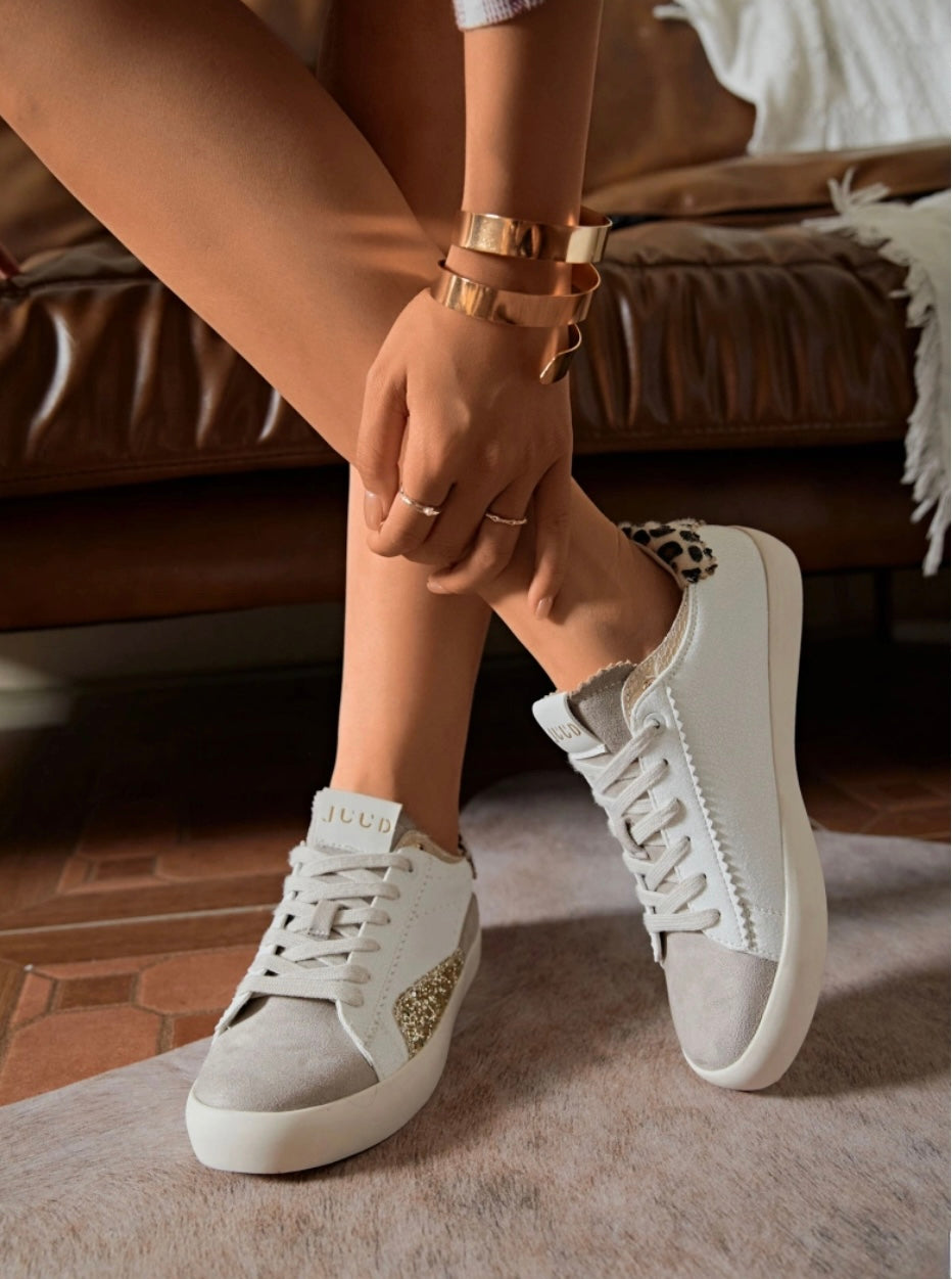A pair of grey and gold leopard print heel trainers with white laces, worn by a person seated on a sofa.