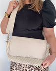 A woman holding a cream and taupe woven zig zag crossbody bag with a foldover feature and a detachable strap.