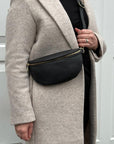 A woman wearing a beige coat is holding a black leather waist bag with a gold-colored zipper.