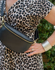 A person wearing a leopard print dress carrying a black leather waist bag with gold hardware.