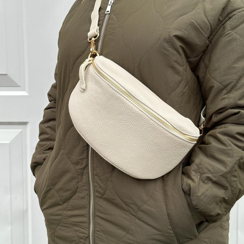 a woman wearing a large cream crossbody/ waist bag with and adjustable strap and gold hardware.
