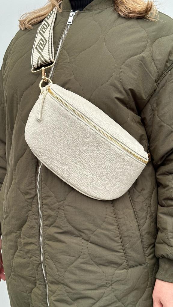 a woman wearing a large cream crossbody/ waist bag with and adjustable strap and gold hardware.
